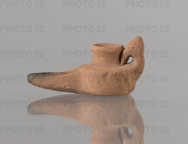 Oil lamp, Islamic art, Arabic culture, Fatimid dynasty 909–1171, Arabic pottery, Egyptian pottery, utility pottery, oil lamp with traces of use, everyday life in the Middle East, 12th century, Egypt, height 3.4 cm, width 9.9 cm, depth 5.0 cm, A small clay object in shades of brown and tan sits against a pale gray background, shaped with a rounded body, a short cylindrical neck opening, and a tapered spout that darkens toward its tip; a curved handle with an oval hole rises from the rear, and the object casts a muted reflection on the glossy surface below, showing variations in texture and small rough spots on the clay.