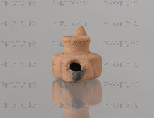 Oil lamp, Fatimid dynasty 909–1171, 12th century, Egypt; Islamic/Arabic pottery; Egyptian utility pottery oil lamp with traces of use; height 3.4 cm, width 9.9 cm, depth 5.0 cm., A small clay object in shades of brown and tan sits centered on a reflective surface, showing a mirrored reflection beneath; the object has a squat, rounded base with a hollow opening at the front that shows darker brown to black discoloration around its rim, and a narrower neck topped by a pointed, conical protrusion, with visible surface irregularities, small cracks, and rough texture against a uniform light gray background.