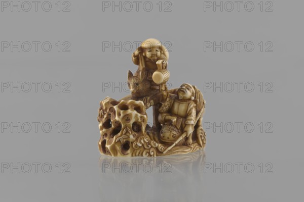 Netsuke in the shape of Daikoku and Ebisu, Ryôji fl. ca. 1886–1900? sculptor, Japanese, Meiji 1868–1912, figurine netsuke katabori, 4th quarter of the 19th century, Japan. Finely carved miniature rocky landscape with a bridge showing Daikoku seated on a rat holding a hammer in his left hand and a bag on his back, and Ebisu holding a fishing rod and riding a giant fish. Meticulous details include facial features, fish-scale outlines, bamboo leaves on openwork rocks, the rat’s fur, and the rodent’s eyes marked with microscopic inlays of black-dyed bone. Height 3.9 cm; width 4.0 cm., A small carved figurine in shades of beige and brown sits against a gray background and is reflected faintly on a glossy surface; the carving depicts a group of figures including a person holding a rounded object above their head, another seated or leaning figure with rolled clothing details, and an animal with pointed ears; the base of the carving resembles an irregular porous rock with hollows and flowing c...