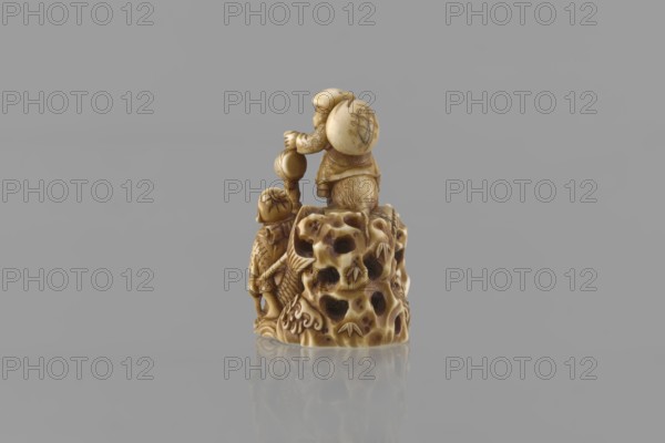 Netsuke in the shape of Daikoku and Ebisu, Ryôji fl. ca. 1886–1900? sculptor, Japanese, Meiji 1868–1912, figurine netsuke katabori. From the group of seven gods of all happiness come two figures, finely carved in a miniature rocky landscape with a bridge: Daikoku, sitting on a rat and holding his attribute—a hammer—in his left hand, with a bag on his back; and Ebisu holding a fishing rod and riding a giant fish. The miniature 4 cm high shows meticulous detail: facial features, fish-scale outlines, bamboo leaves on the openwork rocks, the rat's fur, and the rodent's eyes marked with microscopic inlays of black-dyed bone. 4th quarter of the 19th century, Japan. Height 3.9 cm; width 4.0 cm., A small carved figurine in shades of beige and brown sits centered on a smooth gray surface with a gray background, showing a group of two figures standing on an irregular, porous base that resembles a textured rock; the taller figure wears a rounded hat and a rounded backpack-like object with carved ...