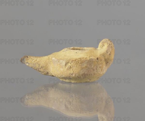 Oil lamp, Islamic art, Islamic culture, Arabic culture, utility ceramics, Arabic ceramics, everyday life in the Middle East, Egyptian ceramics, 8th–9th century, Egypt place of origin, oil lamp, height 9.4 cm, width 5.7 cm, depth 4.1 cm, A roughly shaped object with a textured, worn surface rests on a reflective surface, showing a mirrored reflection below. The object is mostly yellow with patches of pale beige and muted brown, and small areas of darker grayish-brown near the narrower end. Its form includes a rounded, bulbous section that curves upward at one side and tapers to a pointed tip at the opposite side. The background is a uniform gray.