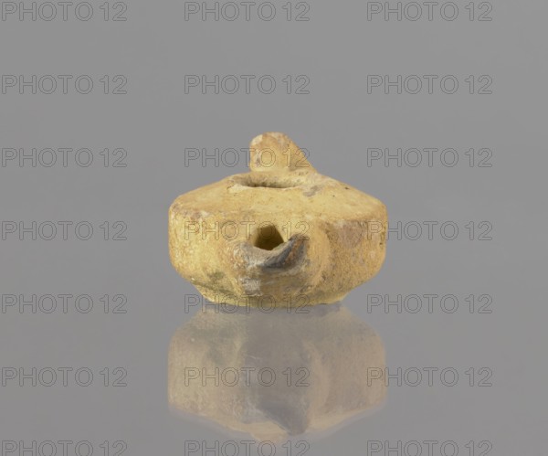 Oil lamp, oil lamp, utility ceramics, Arabic ceramics, Egyptian ceramics, Islamic art, Islamic culture, Arabic culture, everyday life in the Middle East, 8th–9th century, Egypt place of origin, height 9.4 cm, width 5.7 cm, depth 4.1 cm, A small, rounded clay object with an irregular, weathered surface in shades of yellow, beige, tan, and hints of brown sits against a smooth gray background; the object has a shallow central depression, a protruding spout-like feature with a darker grayish-brown interior, and a small raised knob on top, and its underside and front are visible along with a muted reflection below it showing similar yellow and beige tones.