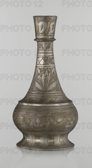Bottle, Islamic art, Arabic culture, geometric and plant motifs, neck slender toward the top ending with a straight spout. Engraved patterns painted with golden-brown paint. Early 20th century, Middle East, vessel, height 21.6 cm, width 10.5 cm., An ornate metal vessel with a narrow neck and flared rim sits upright against a plain light gray background; the vessel displays an overall metallic silver-gray color with areas of darker gray and subtle brownish or bronze tones, and its surface is covered in engraved decorative patterns including leaf-like and curved motifs arranged in bands around the neck, shoulder, and bulbous body, with linear ridges and horizontal bands separating the patterned sections; the base flares outward and shows vertical engraved lines and a slightly reflective surface that creates a faint mirrored reflection on the surface beneath.