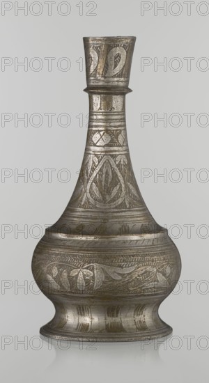 Bottle, Islamic art, Arabic, geometric ornament and plant motifs. Neck slender toward the top ending with a straight spout. Engraved patterns painted with golden-brown paint. Early 20th century, Middle East. Vessel; height 21.6 cm, width 10.5 cm., A single metallic vessel with a tall narrow neck that flares into a wider bulbous body and then narrows to a low pedestal base is shown against a plain light gray background; the vessel surface displays engraved decorative patterns of leaves and geometric shapes, with areas of darker patina and lighter metallic highlights creating a mottled silver and bronze appearance.