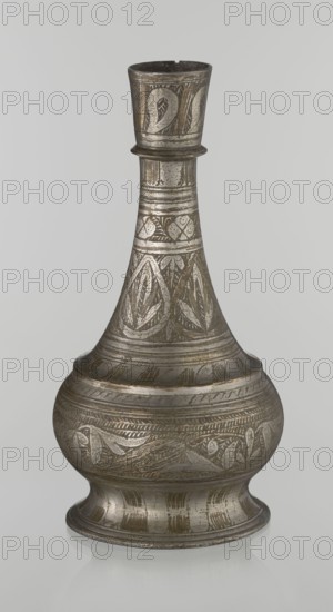 Bottle, Islamic art, Arabic culture, geometric ornament, plant motifs; neck slender toward the top ending with a straight spout; engraved patterns painted with golden-brown paint; early 20th century, Middle East; vessel; height 21.6 cm, width 10.5 cm, The image shows a single metal vessel with a bulbous base, a narrowing neck, and a flared rim, standing against a plain light gray background. The vessel has engraved and incised decorative patterns covering most surfaces, including repeating leaf-like and geometric motifs. The metal surface appears aged with a patina, showing shades of silver-gray and darker gray, along with areas of brownish or bronze discoloration. The reflections and subtle highlights emphasize the metallic texture and the engraved details.