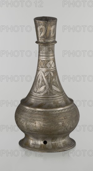 Bottle, Islamic art, Arabic, geometric ornament, plant motifs; neck slender toward the top ending with a straight spout; engraved patterns painted with golden-brown paint; early 20th century, Middle East; vessel; height 21.6 cm, width 10.5 cm., An antique metal vessel with a narrow neck and bulbous body sits against a plain light gray background; the surface shows engraved decorative patterns including leaf-like motifs and bands of etched lines, with areas of darker tarnish and lighter silver highlights giving overall colors of gray, silver, and muted brownish tones; the vessel has a flared rim, a rounded base with a small round hole, and an overall aged, slightly reflective finish.