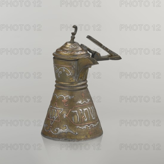 Coffee pot, gift, Arabic and Jewish cultural influences; cezve coffee vessel, beginning of the 20th century, Palestine. Decoration: Hebrew inscriptions inlaid in copper, Star of Solomon motif six-pointed star, arabesque patterns, crescent symbol. Hinged lid hinge halfway down the handle; lid topped with a crescent. Inscriptions deciphered: words including drinking; in the hexagonal star Gan Eden, garden; in the cartouche for celebration; under the handle, in the Seal of Solomon/Star of David Mim from the word water. Vessel: height 15.5 cm, width 15.5 cm, diameter 8.7 cm., A metal pitcher with a hinged lid and a curved spout sits against a plain light gray background; the pitcher has a flared conical base and a narrower neck with decorative raised bands, a small ornate finial on the lid, and a simple lever handle attached near the top. The surface shows a mix of metallic colors including brassy gold, darker bronze, and accents of silver and copper, with areas of darker patina and wear. ...