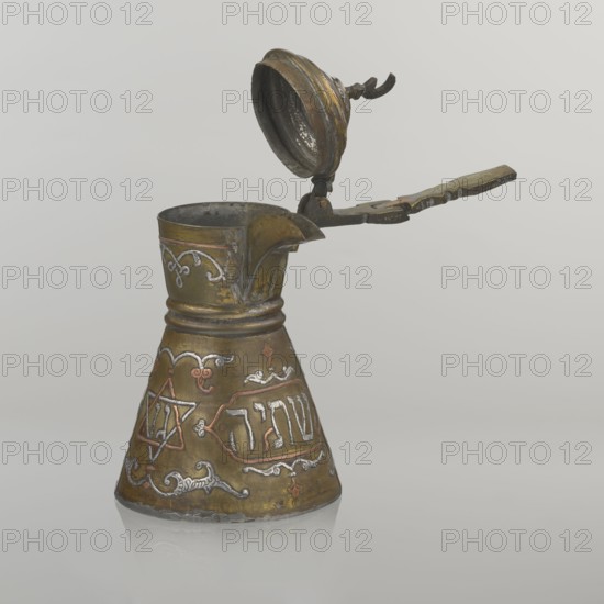 Coffee pot, gift; Arabic and Jewish cultural influences; beverage vessel cezve, beginning of the 20th century, Palestine. Materials/decoration: copper with Hebrew inscriptions inlaid in copper, Star of Solomon six-pointed star motif, arabesque patterns; hinged lid hinge halfway down the handle topped with a crescent. Inscriptions deciphered: words including drinking, in the hexagonal star Gan Eden, garden, in the cartouche for celebration, and under the handle in the Star of Solomon seal Mim from the word water. Dimensions: height 15.5 cm, width 15.5 cm, diameter 8.7 cm., An ornate metal jug with a hinged lid and handle sits against a light gray background; the jug has a flared conical base narrowing toward the neck, a pointed spout, and a top lid that is open and attached to a long, decorative handle with a twisted element near the hinge. The surface shows decorative raised patterns and embossed motifs in contrasting metallic tones, with visible areas of brass-like gold, darker bronze...