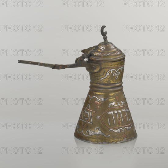 Coffee pot, Arabic and Jewish cultural influences, beverages, cezve vessel. Decoration: Hebrew inscriptions inlaid in copper; Star of Solomon motif; arabesque patterns. Hinged lid with hinge halfway down the handle; lid topped with a crescent. Inscriptions deciphered: readings include drinking; in the hexagonal star Gan Eden, garden; in the cartouche for celebration; under the handle, in the Seal of Solomon/Star of David Mim from the word water. Early 20th century, Palestine. Vessel: height 15.5 cm, width 15.5 cm, diameter 8.7 cm., A small metal vessel with a conical base and a hinged lid attached to a long thin handle is shown against a pale background, the object displaying a worn metallic patina in shades of brass, gold, brown, and dark gray with areas of silver and copper-colored inlay; the surface is decorated with ornate curving motifs and a star-shaped design, and the lid is topped with a small decorative finial while the hinged mechanism and handle appear darker and slightly ru...