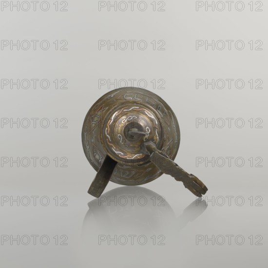 Coffee pot cezve, beginning of the 20th century, Palestine. Copper vessel, height 15.5 cm, width 15.5 cm, diameter 8.7 cm. Arabic and Jewish cultural influences; used for coffee. Decoration: Hebrew inscriptions inlaid in copper, Star of Solomon motif six-pointed star, arabesque patterns; hinged lid with hinge halfway down the handle and lid topped with a crescent. Inscriptions deciphered: hexagonal star contains Gan Eden, garden; cartouche reads for celebration; under the handle, in the Star of Solomon Mim from the word water; also the word drinking., A small metallic object with a rounded, dome-like top sits on a reflective surface, showing a muted palette of brown, bronze, gold, and hints of silver and copper; the surface of the object displays engraved or embossed decorative patterns and sinuous lines in lighter metallic tones, and a short attached arm or lever extends from the side with an irregular, slightly tapered shape and a darker, aged brown finish, while the background is a ...