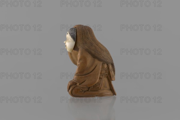Netsuke in the shape of a nô theater actor wearing an Okame mask, Hôjitsu fl. ca. 1800–1860 sculptor. Netsuke in the form of a figurine of a sitting nô theater actor wearing an Okame mask. The entire figure is carved in wood; the mask is made of ivory, carved, engraved, and stained with black ink. The kimono is girded with an obi belt. Okame is depicted as a chubby, pleasant, smiling woman. Signature on the underside: Hojitsu. 19th century Edo period, 1603–1868, Japan. Sculpture; height 4.0 cm, width 2.6 cm., A small carved figure is shown in profile against a plain gray background, depicting a person kneeling with long textured hair flowing down the back and wearing a layered robe with carved geometric patterns on the skirt and back; the face is a lighter smooth material with visible hairline details and a neutral expression, and the overall tones visible are shades of brown, tan, cream, gray, and a little black in the hair detailing.