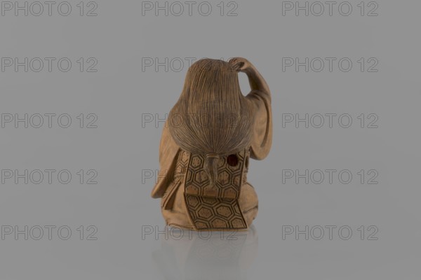 Netsuke in the shape of a nô theater actor wearing an Okame mask, Hôjitsu fl. ca. 1800–1860 sculptor. Netsuke in the form of a figurine of a sitting nô theater actor wearing an Okame mask. The entire figure is carved in wood; the mask is ivory, carved, engraved, and stained with black ink. The kimono is girded with an obi belt. Okame is depicted as a chubby, pleasant, smiling woman. Signature on the underside: Hojitsu. 19th century Edo period, 1603–1868, Japan. Sculpture; height 4.0 cm, width 2.6 cm., A small carved wooden figurine shown from the back with long textured hair tied at the nape, wearing a patterned garment with hexagonal motifs and a flap extending downward, one arm raised and bent at the elbow, the surface showing carved details and a smooth finish; the scene includes a reflective surface beneath the figurine and a plain gray background, with visible colors of brown and gray.