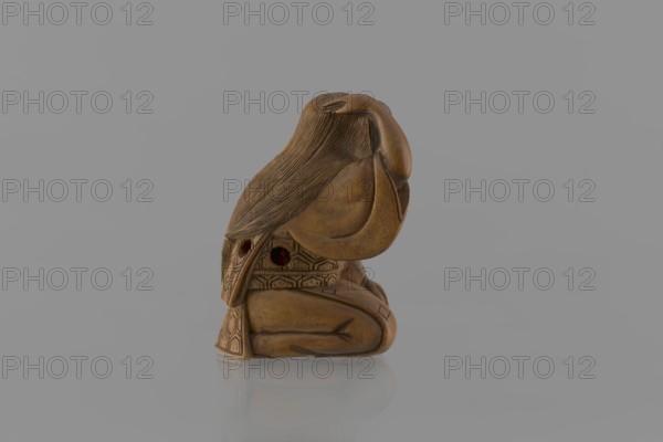 Netsuke in the shape of a nô theater actor wearing an Okame mask, Hôjitsu fl. ca. 1800–1860 sculptor. Netsuke in the form of a figurine of a sitting nô theater actor wearing an Okame mask. The entire figure is carved in wood; the mask is made of ivory, carved, engraved, and stained with black ink. The kimono is girded with an obi belt. The goddess Okame is depicted as a chubby, pleasant, smiling woman. Signature on the underside: Hojitsu. 19th century Edo period, 1603–1868, Japan. Sculpture. Height 4.0 cm, width 2.6 cm., A small carved wooden figure in brown tones sits on a reflective light gray surface against a soft gray background; the figure appears to be in a seated, curled posture with long textured hair flowing over its back and an arm raised near its head, and the carved surface shows patterned geometric details and smoothly rounded shapes with subtle darker brown shading.