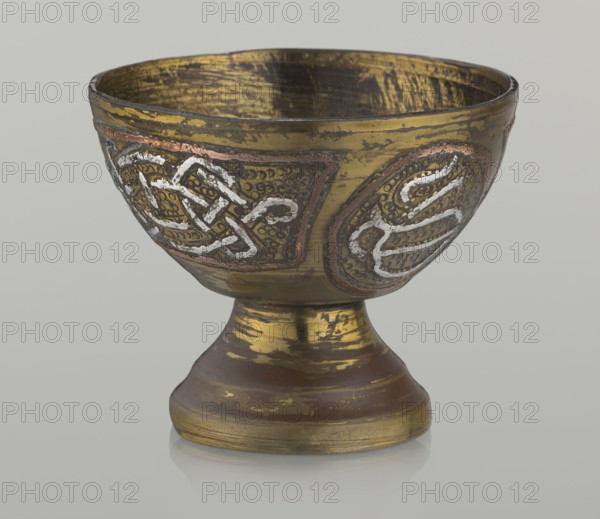 Bowl, decorated with braid and stylized Arabic inscriptions in cartouches on a punched background; Arabic inscriptions and calligraphy; geometric motifs; early 20th century; Middle East, possibly Palestine; vessel; height 4.7 cm, diameter 5.5 cm., A metal chalice with a rounded bowl atop a flared stem and circular base sits against a neutral background; the surface shows worn gilding in gold and brown tones with areas of darker tarnish and a few patches of reddish metal; the bowl features several raised decorative panels with textured backgrounds and braided linear motifs in silvery white and coppery outlines; the interior of the bowl shows a distressed gold finish with visible brushlike marks; the base and stem display bands of rubbed gold and brown creating a weathered appearance.