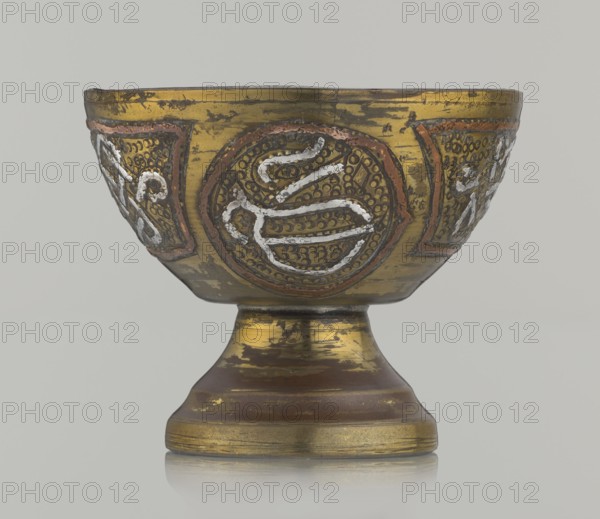 Bowl, Decoration with braid and stylized Arabic inscriptions in cartouches; punched background. Arabic inscriptions; calligraphy; geometric motifs. Early 20th century. Middle East, possibly Palestine. Vessel; height 4.7 cm, diameter 5.5 cm., A decorative metal bowl on a pedestal with a rounded basin and flared foot, showing ornate raised circular medallions and panel decorations around the exterior. The surface displays worn gilding with visible patches of exposed darker metal and areas of brownish patina. The decorative motifs include raised textured dots and curving raised lines highlighted in a silvery-white inlay and bordered by a reddish-brown trim. The foot and lower stem show horizontal banding and worn gold areas over a darker brown base. The background is a neutral pale gray.