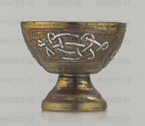 Bowl, gift, Islamic art, Arabic culture, inscriptions, Arabic calligraphy, geometric motifs. Decoration with braid and stylized Arabic inscriptions in cartouches; punched background. Early 20th century, Middle East, possibly Palestine. Vessel; height 4.7 cm, diameter 5.5 cm., A metal goblet shown from the front with a rounded bowl atop a flared pedestal base, featuring decorative embossed patterns and raised interlaced designs. The surface shows a worn metallic finish with visible patches and streaks. Colors visible include gold, bronze, brown, copper, silver, and a pale gray background. The bowl has areas of textured dotted decoration and triangular panels containing the raised interlaced motif, and the base displays bands of rubbed metallic tones and reflective highlights.