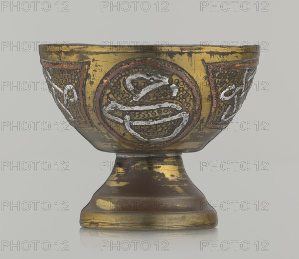 Bowl, early 20th century, Palestine, metal vessel, height 4.7 cm, diameter 5.5 cm. Islamic art; Arabic inscriptions and calligraphy; decoration with braid and stylized Arabic inscriptions in cartouches on a punched background; geometric motifs., A shallow bowl on a pedestal with a rounded foot, shown against a plain light gray background, featuring a metallic surface with visible wear and patches where the finish has come off; the bowl displays a predominant gold color with areas of darker brown and muted copper tones, and raised decorative circular motifs with textured dot backgrounds and raised irregular white-silver lines, framed by reddish-brown borders, with the pedestal showing horizontal bands of gold and brown and patches of worn gold revealing underlying darker metal.