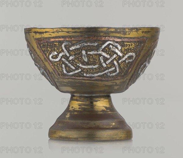 Bowl, early 20th century, Palestine possibly, ceramic vessel decorated with braid and stylized Arabic inscriptions in cartouches, punched background; Islamic art Arabic culture, Arabic calligraphic inscriptions; height 4.7 cm, diameter 5.5 cm., An ornate metal bowl on a pedestal with a worn metallic surface showing gold, bronze, copper, and silver tones, decorated with an embossed interlacing knot pattern highlighted in silver on a textured gold background framed by copper accents, with areas of darker brown patina and patches where the gold finish is rubbed away, all set against a pale gray background.