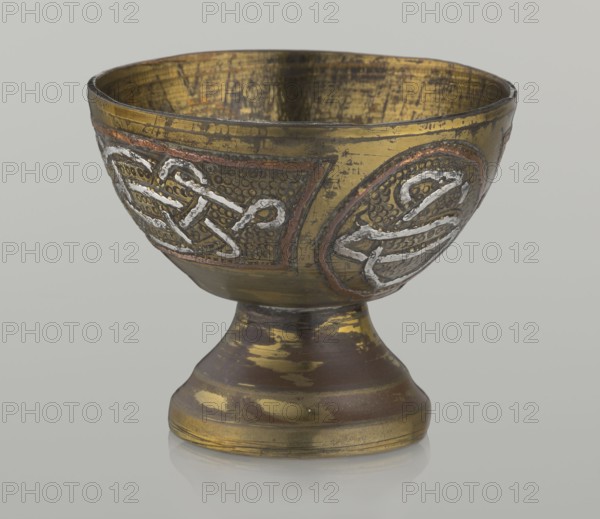 Bowl, early 20th century, Middle East possibly Palestine. Vessel, height 4.7 cm, diameter 5.5 cm. Decoration with braid and stylized Arabic inscriptions in cartouches; punched background. Islamic art; Arabic inscriptions; calligraphy; geometric motifs., A metal chalice sits on a short pedestal with a rounded base, showing a worn metallic surface with gold, brown, and darker bronze tones; the bowl features decorative raised designs outlined in a silver color against textured dark backgrounds with traces of reddish accents, and the interior of the bowl displays worn gold and dark streaks.