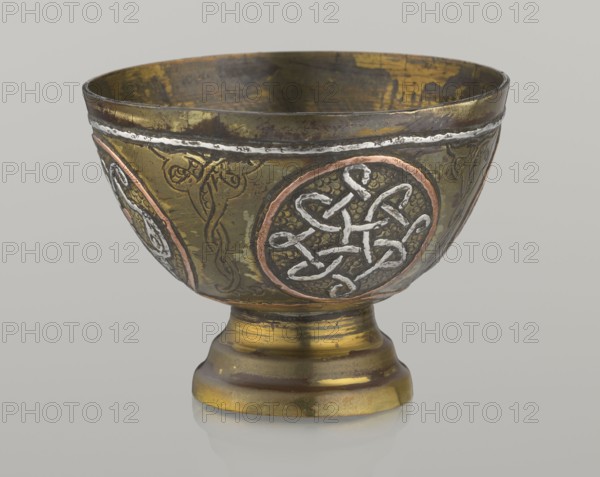 Bowl, gift, Islamic art, Arabic, geometric motifs; decorated with braided and arabesque motifs in cartouches; background punched. 20th century, Middle East, possibly Palestine. Vessel; height 4.3 cm, diameter 5.8 cm., A metal chalice with a rounded bowl and pedestal base, showing a worn metallic surface in gold, brass, and brown tones with areas of darker patina; the bowl exterior features circular medallions with raised, intertwined line decorations in silver and copper hues set against textured dark backgrounds, separated by engraved vine-like motifs in dark brown and gold; the rim shows a thin band of silvery metal and a darker interior with gold and brown patches; the pedestal displays horizontal ridges and a mix of gold, bronze, and brown finishes, and the whole object sits on a reflective grey surface against a plain light grey background.