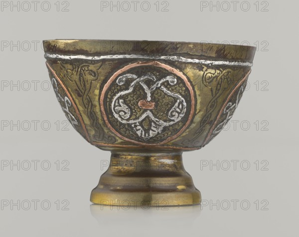 Bowl;, 20th century; Middle East, possibly Palestine; Islamic/Arabic; vessel, height 4.3 cm, diameter 5.8 cm; decorated with braided and arabesque motifs in cartouches on a punched background., A metallic bowl on a pedestal with decorative circular medallions and ornate patterns around its exterior, showing a mix of metallic gold, brass, copper, silver, and dark brown tones with areas of wear and patina; the bowl has raised and engraved motifs in contrasting metals, a flared rim with a thin silver-colored band near the top, and a rounded foot with horizontal stepped profiles, all set against a plain light gray background.