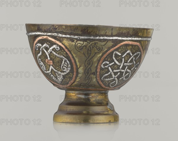 Bowl, Islamic art, Arabic culture; 20th century, possibly Palestine; vessel, height 4.3 cm, diameter 5.8 cm; decoration with braided and arabesque motifs in cartouches, background punched., A metal goblet sits against a plain light gray background, showing a rounded bowl on a short flared stem and circular base. The surface of the goblet displays a metallic olive-green patina with areas of darker brown and worn gold tones, and there are copper-colored rings around two oval medallions on the bowl. Each medallion contains raised decorative knotwork rendered in a bright silvery-white metal, contrasted against a textured dark background. A thin silvery band runs near the rim, and faint engraved lines and darker linear markings appear across the body. The base shows a mix of gold, brown, and greenish hues with patches of darker wear and a small area of exposed lighter metal. Overall the palette includes green, brown, gold, copper, silver, white, and dark gray.