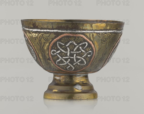 Bowl, Islamic art, Arabic; Decoration with braided and arabesque motifs in cartouches. Background punched. 20th century, Middle East, possibly Palestine. Vessel; height 4.3 cm, diameter 5.8 cm., A metal goblet with a rounded bowl on a short pedestal base, showing a patina of greenish-gold, brown, and dark bronze tones, with areas of worn yellow-gold and small patches of silvery metal. The bowl surface is decorated with circular medallion designs outlined in a coppery reddish tone and filled with silvery raised knotwork against a darker textured background, and there are linear braided and vine-like engraved patterns in darker and brown hues. The rim has a thin silvery band and the pedestal shows wear with greenish-gold, brown, and darker brown bands and a noticeable silvery blemish, all set against a plain pale gray background.