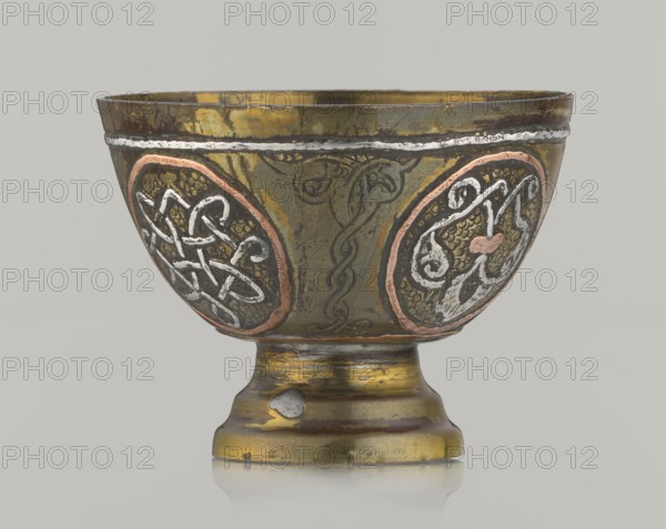 Bowl, Islamic art, Arabic; 20th century, Middle East, possibly Palestine; vessel, height 4.3 cm, diameter 5.8 cm. Decoration with braided and arabesque motifs in cartouches; background punched., A metal goblet with a rounded bowl and a short pedestal base, showing a mottled surface in brass tones of gold and olive with areas of darker brown and green patina; the bowl is decorated with oval medallions outlined in a coppery pink and filled with textured backgrounds and raised swirling motifs in silvery white and darker metal tones, and a raised wavy silver band runs near the rim; the stem and base display worn gold and bronze coloration with small spots where the surface has been rubbed to a lighter silvery tone; the overall palette includes gold, olive, brown, green, coppery pink, and silvery white.