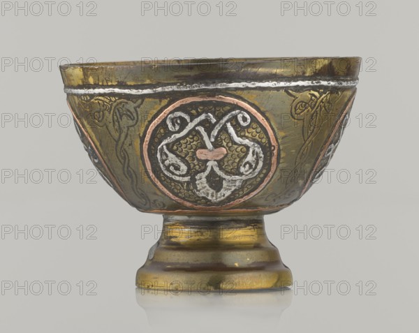 Bowl, gift, Islamic art, Arabic, geometric motifs. Decoration with braided and arabesque motifs in cartouches; background punched. 20th century, Middle East, possibly Palestine. Vessel; height 4.3 cm, diameter 5.8 cm., A metal bowl with a short pedestal base sits against a plain light gray background; the bowl shows a mix of metallic colors including gold, brass, copper, silver, and dark gray, with areas of patina and wear. The body of the bowl features decorative circular medallions with raised, curving motifs in contrasting metallic tones and a textured background inside the medallions. Around the bowl there are slender ribbon-like engraved designs and narrow bands near the rim highlighted in a lighter metallic color. The pedestal has horizontal wear marks and layered metallic hues, and the whole object casts a faint reflection on the surface beneath it.
