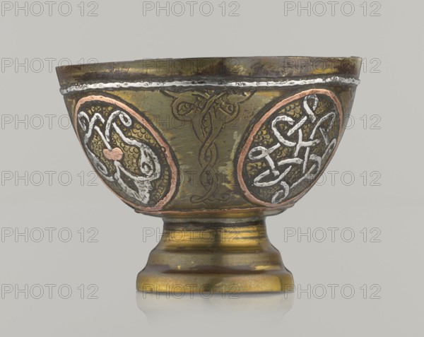 Bowl, Islamic art, Arabic culture, geometric motifs, decoration with braided and arabesque motifs in cartouches; background punched. 20th century, Middle East, possibly Palestine. Vessel; height 4.3 cm, diameter 5.8 cm., A decorative metal bowl on a short pedestal base with a rounded foot, shown against a plain light gray background. The bowl displays a mottled greenish-brass surface with areas of darker tarnish and patina. Around the exterior are a few circular medallions outlined in a coppery pink tone containing raised interlaced designs highlighted in a silvery white. There are also engraved curving motifs between the medallions in a darker brownish-green. The rim has a thin silvery band with uneven texture, and the pedestal shows bands of golden brass, darker brown, and muted green. Overall visible colors include greenish-brass, brown, coppery pink, silvery white, dark brown, gold, and light gray.