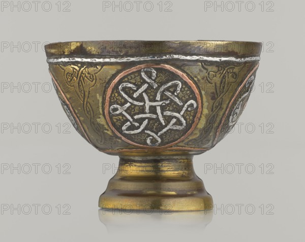 Bowl, gift, Islamic art, Arabic culture, geometric motifs; decoration with braided and arabesque motifs in cartouches; background punched. 20th century, Middle East, possibly Palestine. Vessel; height 4.3 cm, diameter 5.8 cm., A metal bowl on a pedestal with a rounded foot and flared rim, showing decorative circular medallions and engraved vine-like motifs; the surface displays a combination of metallic gold, dark brown, copper, and silver tones, with textured areas and polished highlights; the background is a plain light gray.