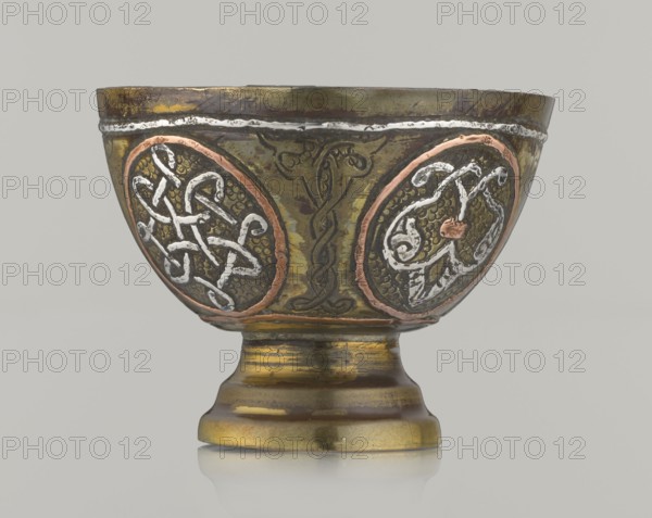 Bowl, 20th century, possibly Palestine, Middle East; Islamic/Arabic art. Vessel, height 4.3 cm, diameter 5.8 cm. Decoration with braided and arabesque motifs in cartouches; background punched; geometric motifs., A decorative metal bowl on a pedestal base with a rounded body and flared rim, showing a patinated surface in shades of olive green and brass yellow with darker brown areas. The bowl features a few circular medallion panels around its body outlined in a coppery rose tone, each filled with textured dark backgrounds and raised interlaced decorative motifs in silvery white. A thin silver-colored band runs near the rim, and there are darker engraved linear designs between the medallions. The pedestal has horizontal stepped rings and shows the same brass yellow and brown coloration with some reflective highlights. The overall palette includes olive green, brass yellow, brown, coppery rose, dark gray, and silvery white.
