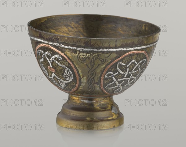 Bowl, Islamic art, Arabic culture, geometric motifs; decoration with braided and arabesque motifs in cartouches; background punched. 20th century, Middle East, possibly Palestine; vessel, height 4.3 cm, diameter 5.8 cm., A metal bowl with a rounded body and a short pedestal base, shown against a light gray background; the bowl surface is primarily brassy yellow with areas of darker brown patina and worn greenish tones, with decorative circular panels on the sides bordered in a coppery pink and filled with raised silver-white intertwined motifs; a silver-white band runs near the rim, and there are engraved dark linear patterns between the panels; the interior shows darker brown and muted gold coloration with visible wear.