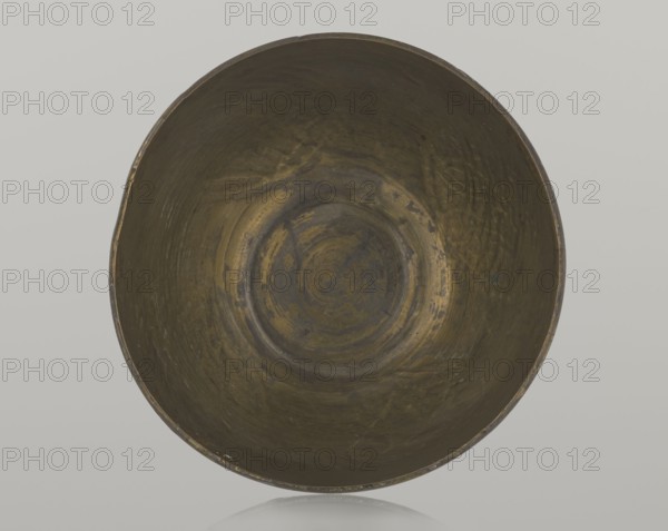 Bowl, Islamic art, Arabic, geometric motifs; decoration with braided and arabesque motifs in cartouches; background punched. 20th century, Middle East, possibly Palestine; vessel, height 4.3 cm, diameter 5.8 cm, A round, shallow bowl viewed from above with a worn, metallic surface showing a mix of dark brown, olive green, and muted gold tones, visible circular striations and irregular patina marks across the interior, a slightly uneven rim with small areas of lighter wear, and a pale gray background.