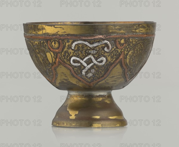 Bowl, gift, Arabic, arabesque ornament, geometric motifs, punched background, 20th century, Middle East, possibly Palestine, vessel; height 4.3 cm, diameter 5.6 cm, A decorative metal bowl on a pedestal with a rounded form, showing a few areas of worn gold and brass tones, a group of darker brown and black patches, and accents of reddish copper; the bowl's body features raised ornamental patterns and a central looping motif in a lighter silvery color set against textured background, with the pedestal reflecting similar gold and brown wear and a smooth curved silhouette, all shown against a plain pale gray background.