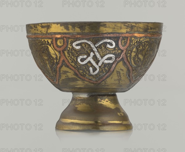 Bowl, gift, Islamic art, Arabic culture, arabesque ornament, geometric motifs, punched background, 20th century, Middle East, possibly Palestine, vessel; height 4.3 cm, diameter 5.6 cm., A decorative chalice-like bowl sits on a rounded foot, showing a metallic surface with worn areas and a patina. The bowl features embossed and engraved patterns around its exterior, including curving, interlaced motifs highlighted in a lighter metallic tone and bordered with a darker outline. There are raised linear decorations in a reddish-copper color tracing triangular panels around the body, and the background of the panels has textured dotwork and engraved designs in darker shades. The overall colors visible are gold, bronze, copper, dark brown, and a lighter silvery-white accent on the central motif, set against a pale neutral background.