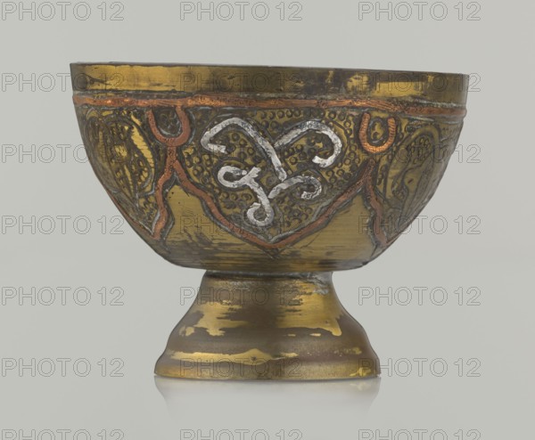 Bowl, 20th century, Middle East possibly Palestine. Ceramic vessel with arabesque and geometric motifs and punched background; height 4.3 cm, diameter 5.6 cm., A decorative metal bowl on a pedestal with an aged, reflective surface showing a mix of gold, brass, copper, and silver tones; the bowl exterior features raised, curving ornamental patterns highlighted in copper and silver against a darker gold background, with textured dotted areas and stylized motifs around the rim; the pedestal base shows worn patches of gold and darker bronze, with irregular areas of patina and surface wear; the overall color palette includes gold, brass, copper, silver, bronze, and muted gray, set against a pale neutral backdrop.