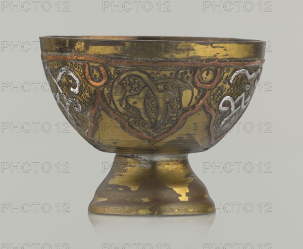 Bowl, Islamic art; Arabic; 20th century, possibly Palestine; vessel with arabesque and geometric motifs, punched background; height 4.3 cm, diameter 5.6 cm., A metal bowl on a short pedestal base with decorative raised patterns and inlaid accents, showing worn and scratched surfaces. The bowl and base are primarily gold and brass tones with patches of darker brownish areas where the finish is worn. There are raised decorative lines and motifs in coppery orange and silvery white against the gold background, with some darker engraved details creating texture. The interior rim and top edge show darker brass coloration, and the overall finish has an aged, patinated look with irregular areas of shine and dullness. The background behind the object is a neutral light gray.
