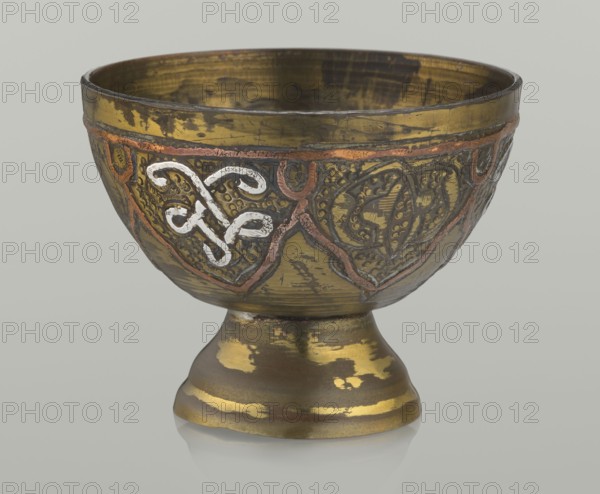 Bowl, Islamic art, Arabic, arabesque ornament, geometric motifs, punched background. 20th century, Middle East, possibly Palestine. Vessel; height 4.3 cm, diameter 5.6 cm., A metal bowl on a short pedestal with a rounded foot, showing worn gold and bronze tones with patches of darker brown and areas where the finish is rubbed away; the bowl exterior has an embossed decorative band with raised curving vine-like motifs highlighted in a silvery white and bordered by a reddish copper-colored outline, set against textured dark brown and black backgrounds; the interior shows similar gold and bronze wear with dark streaks; the background behind the object is a pale neutral gray.