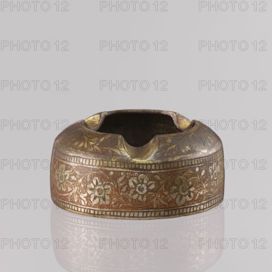 Ashtray, applied art; floral tendril decoration on the sides; composition with a botanical pattern and two branches on the flattened part of the wall; engraved patterns colored with orange and black pastes; plant, geometric, leaf and flower ornaments; tobacco smoking accessory; 1st half of the 20th century; North India; height 3.2 cm, diameter 7.0 cm., A round metal object with a shallow central cavity and several curved notches along its rim, decorated with an ornate floral band around its sides; the surface shows metallic gold tones with areas of reddish-brown and darker brown patina, and a few lighter highlights where the metal catches the light.