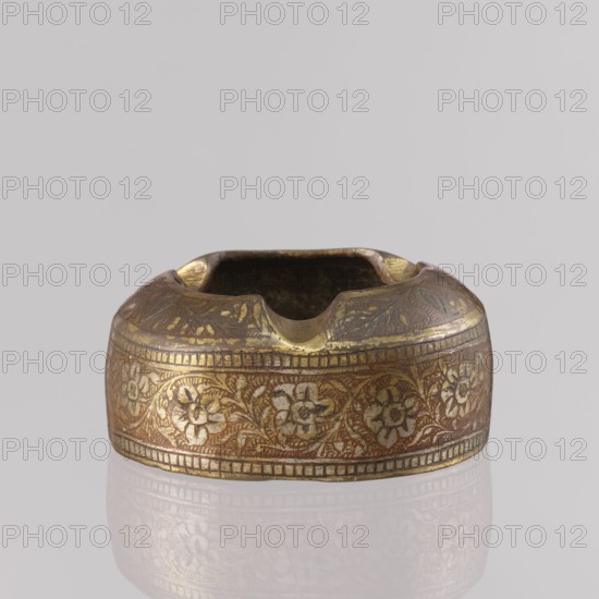 Ashtray; applied art, tobacco smoking accessory. Decoration: floral tendril on the sides; composition with a botanical pattern and two branches on the flattened part of the wall. Engraved patterns colored with orange and black pastes. Plant ornaments, geometric ornaments, leaves, flowers. 1st half of the 20th century. North India. Height 3.2 cm; diameter 7.0 cm. Maker: unknown., A small round metal object with a shallow square opening at the top, showing a few notches along its rim; the surface is decorated with an ornate floral band encircling the body and a patterned border above and below. The visible colors are gold, brown, and reddish tones, with areas of darker patina and lighter worn highlights. The object sits on a reflective pale surface against a light gray background.