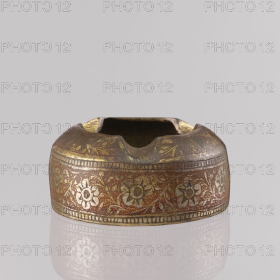 Ashtray; maker, early 1st half of the 20th century; North India. Applied art smoking accessory of Indian culture, used for tobacco smoking. Decoration: floral tendril on the sides; composition with a botanical pattern and two branches on the flattened part of the wall. Engraved patterns colored with orange and black pastes; plant ornaments, geometric ornaments, leaves, flowers. Height 3.2 cm; diameter 7.0 cm., A low, rounded metal object with a shallow rectangular opening and three rounded notches along its rim, decorated around its sides with a repeating floral band; visible colors include gold, reddish-brown, dark brown, and a pale gray background, with worn and darker areas on the rim and inner surfaces.