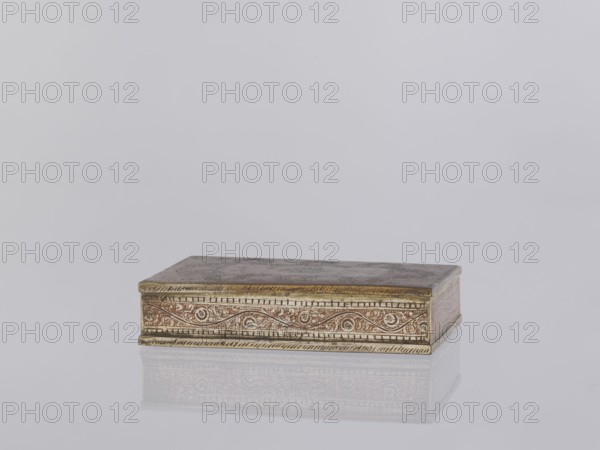 Box with lid. Indian North India. 1st half of the 20th century. Applied art; smoking accessory/casket/box. Decorated on the sides with plant twigs; geometric ornaments. Paste colors: red, orange, black paste pressed into engraved patterns. Height 2.8 cm; width 12.8 cm; depth 9.0 cm., A rectangular ornate box sits on a reflective surface against a pale gray background, showing a metallic appearance with gold and brass tones and areas of reddish-brown patina; the box has decorative engraved patterns along its sides with swirling motifs and border detailing in darker and lighter gold hues, and its top shows a slightly worn, mottled finish with muted brown and grayish areas, the whole object casting a soft reflection on the surface below.