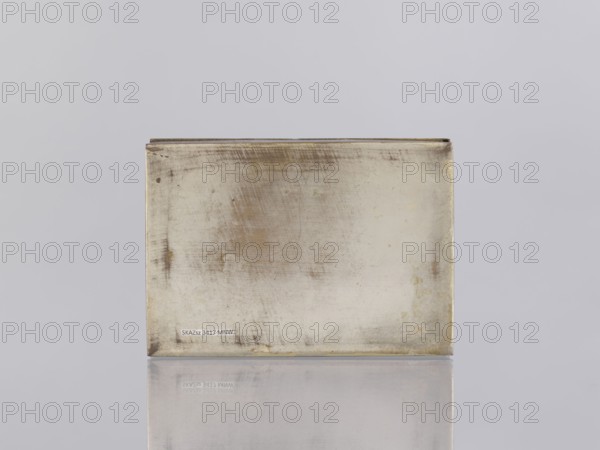 Box with lid. Box decorated on the sides with plant twigs. Paste colors: red, orange, black paste pressed into engraved patterns. 1st half of the 20th century. North India. Indian art; smoking accessories; caskets; boxes; plant ornaments; geometric ornaments; applied art. Height 2.8 cm; width 12.8 cm; depth 9.0 cm., A rectangular metallic box with a worn, tarnished surface sits centered on a reflective surface against a plain light gray background; the box shows areas of dull silver and muted gold tones with darker gray and brownish stains and scratches, and a faint reflection of the box appears below it.