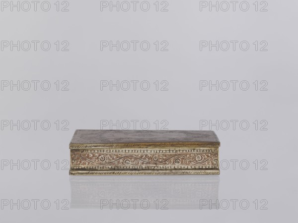 Box with lid. Box decorated on the sides with plant twigs. Paste colors: red, orange, black paste pressed into engraved patterns. Smoking accessory/casket/box; applied art with plant and geometric ornaments. Indian art, North India, 1st half of the 20th century. Height 2.8 cm; width 12.8 cm; depth 9.0 cm., A rectangular decorative box sits centered on a reflective surface, showing a subtle mirrored reflection beneath it; the box has a flat lid and ornate carved or embossed sides with swirling patterns and border detailing, displaying metallic tones of gold and bronze with hints of reddish-brown, and the scene background is a smooth pale gray.