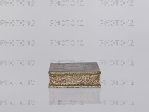 Box with lid. Indian, North India; 1st half of the 20th century. Box decorated on the sides with plant twigs; paste colors: red, orange, black pressed into engraved patterns. Smoking accessory/casket/box; applied art. Materials and ornament: plant and geometric ornaments. Dimensions: height 2.8 cm; width 12.8 cm; depth 9.0 cm., A small rectangular box sits centered on a reflective surface, showing a faint mirrored image beneath it. The box has ornate carved or engraved patterns along its visible front edge and sides, with a decorative border and swirling motifs. The top of the box appears slightly darker with a worn surface. Visible colors include gold tones on the box's decorative borders, brownish and reddish hues within the carved areas, and shades of gray and off-white in the background and reflective surface.