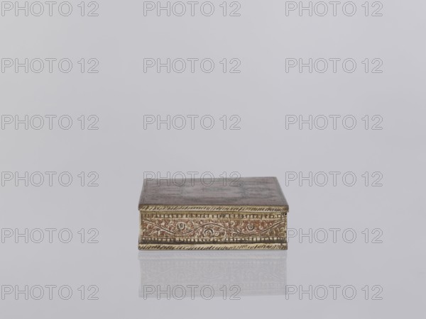 Box with lid. Decorated on the sides with plant twigs. Paste colors: red, orange, black paste pressed into engraved patterns. 1st half of the 20th century. North India. Indian art; smoking accessories; casket/box. Materials/technique: applied art, plant and geometric ornament. Dimensions: height 2.8 cm, width 12.8 cm, depth 9.0 cm., A small rectangular box sits centered on a smooth reflective surface, showing a faint mirrored reflection beneath it; the box has an ornate patterned band along its front edge with etched decorative motifs and a slightly raised border, and the lid appears flat and slightly weathered; visible colors include muted gold, brown, reddish-brown, and hints of gray against a pale light-gray background.