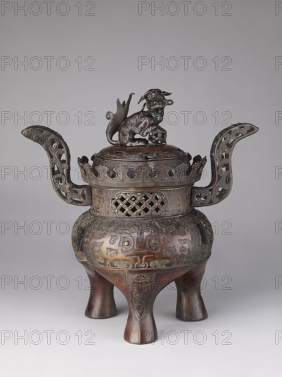 Ding-type incense burner koro with cover, Yoshida maker, karashishi lion/komainu/fo and taotie iconography, meander and plant tendril ornamentation. Incense burner with two openwork handles. Cylindrical neck with a partially openwork lattice pattern terminating in crenellation. Domed cover topped with a fo figurine holding a sphere openwork. Engraved and relief patterns over the entire surface. 19th/20th century Meiji period, Kyoto, Japan. Height 47.0 cm; diameter 29.0 cm., An ornate metal vessel stands on three rounded legs, each leg showing a smooth brown surface with darker brown shading and raised decorative panels. The body of the vessel is bulbous with a carved band of swirling and stylized motifs in brown and darker bronze tones wrapping around it, and a pair of sculpted animal heads in darker metal projecting from the sides. A pierced lattice band appears near the top in darker brown metal, above which a scalloped rim with openwork flourishes encircles the lid. Two high, curved...