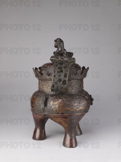 Ding-type incense burner koro with cover, Yoshida maker;, noted. Japanese, Meiji period 1868–1912, 19th/20th century. Cast metal with openwork; cylindrical neck with partially open lattice pattern terminating in crenellation; domed cover topped by a fo karashishi/komainu figurine holding a sphere openwork. Incised and repoussé/relief ornament across the surface including taotie, karashishi/lion motifs, meander borders, and plant tendrils. From Kyoto. Incense burner with two openwork handles. Height 47.0 cm; diameter 29.0 cm., An ornate tripod vessel with a rounded body and three flared legs stands against a plain light gray background; the vessel is predominantly brown with darker brown and black areas and a subtle greenish patina in places, and it is richly decorated with carved and relief patterns all over its surface, including stylized swirling and geometric motifs; a lid with a carved beast finial sits on top, and a tall decorative handle or crest with openwork scrolling designs r...