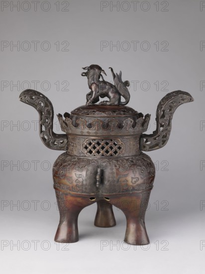 Ding-type incense burner koro with cover, Yoshida maker. Incense burner with two openwork handles. Cylindrical neck with a partially openwork lattice pattern terminating in crenellation. Domed cover topped with a fo karashishi/komainu/guardian lion figurine holding a sphere openwork. Engraved and relief patterns covering the surface, including taotie, meander ornament, and plant tendril motifs. Meiji period, 19th–20th century Japan, Kyoto. Height 47.0 cm; diameter 29.0 cm., An ornate three-legged metal vessel with a dark brown and bronze patina stands against a neutral gray background, featuring a rounded body raised on a group of three flared legs and decorated with intricate relief patterns and carved motifs; a domed lid topped by a sculpted animal figure sits above a pierced collar with latticework, and two elaborately curved handles with openwork scroll designs extend horizontally from either side, all showing aged metallic tones of brown, bronze, and hints of greenish oxidation.